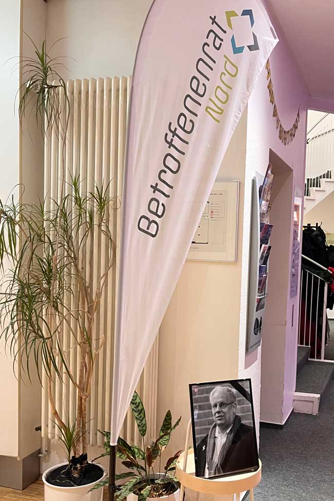 Eine Beachflag mit dem Logo des Betroffenenrats Nord steht hinter einem Foto des verstorbenen Mitglieds Raphael Ohlms